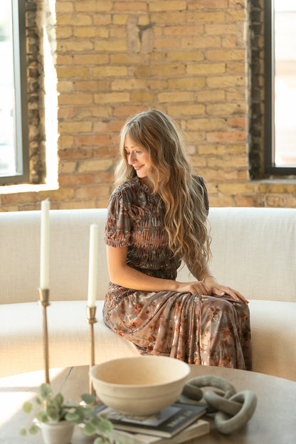 Seated view of muted floral maxi dress showing puff sleeves, smocked bodice, and soft flowing skirt.