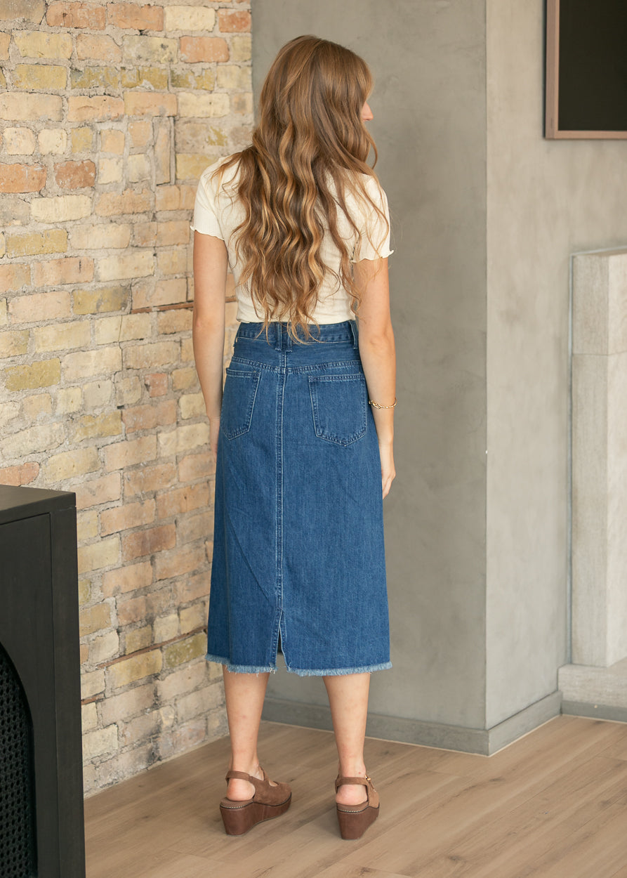 A woman stands with her back to the camera wearing a blue denim midi skirt, a cream short-sleeve top, and brown wedge sandals in an indoor setting with a brick wall and concrete wall background.