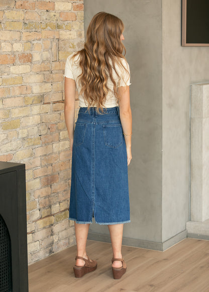 A woman stands with her back to the camera wearing a blue denim midi skirt, a cream short-sleeve top, and brown wedge sandals in an indoor setting with a brick wall and concrete wall background.