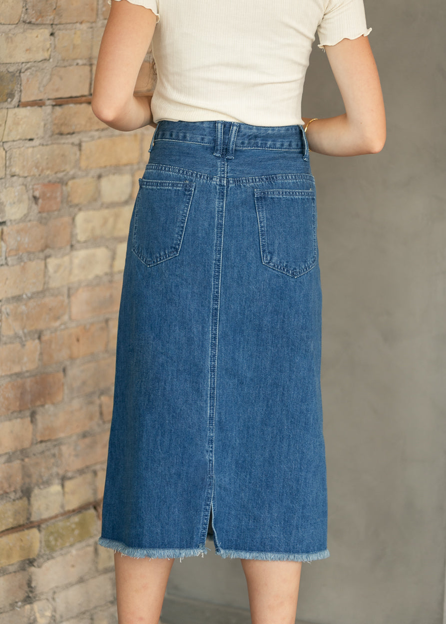 A person wearing a blue denim midi skirt with a front slit and back pockets, paired with a cream short-sleeve top, standing against a brick and concrete wall.