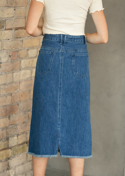 A person wearing a blue denim midi skirt with a front slit and back pockets, paired with a cream short-sleeve top, standing against a brick and concrete wall.