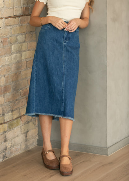 A person wearing a mid-length blue denim skirt with a raw hem, paired with a cream top and brown Mary Jane shoes, standing in a corner with a brick wall, concrete pillar, and wood floor.