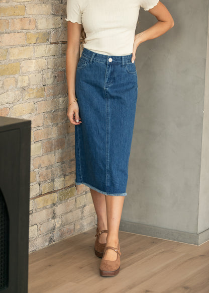 A model wearing a blue denim midi skirt with a raw hem, paired with a cream short-sleeve top and brown Mary Jane flats, standing in front of a brick wall and concrete wall.