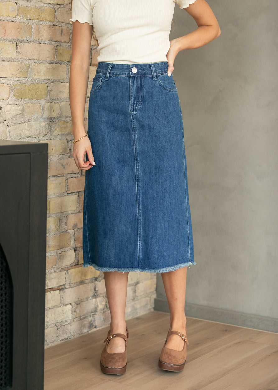 A woman wearing a mid-length blue denim skirt with a raw hem, paired with a cream short-sleeve top and brown Mary Jane shoes, standing in front of a brick wall and concrete wall.