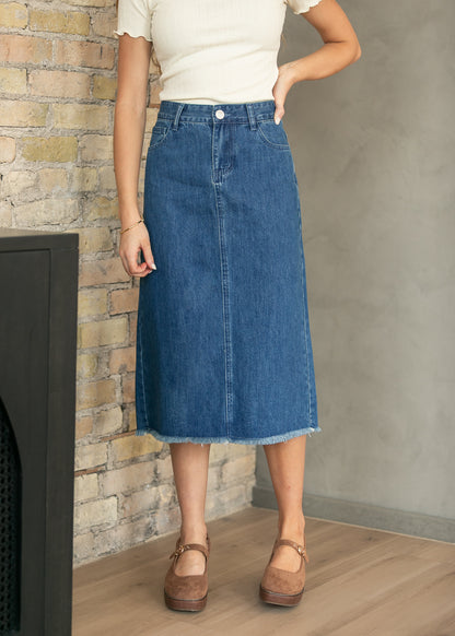 A woman wearing a mid-length blue denim skirt with a raw hem, paired with a cream short-sleeve top and brown Mary Jane shoes, standing in front of a brick wall and concrete wall.