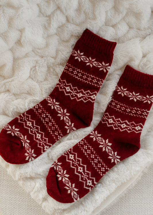 AK red knit crew socks with cream Fair Isle snowflake design and ribbed cuffs, laid on a soft ivory blanket.