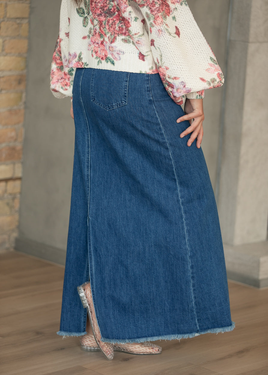 Medium-wash denim maxi skirt from back with long slit and raw-edge hem, styled with a floral sweater.