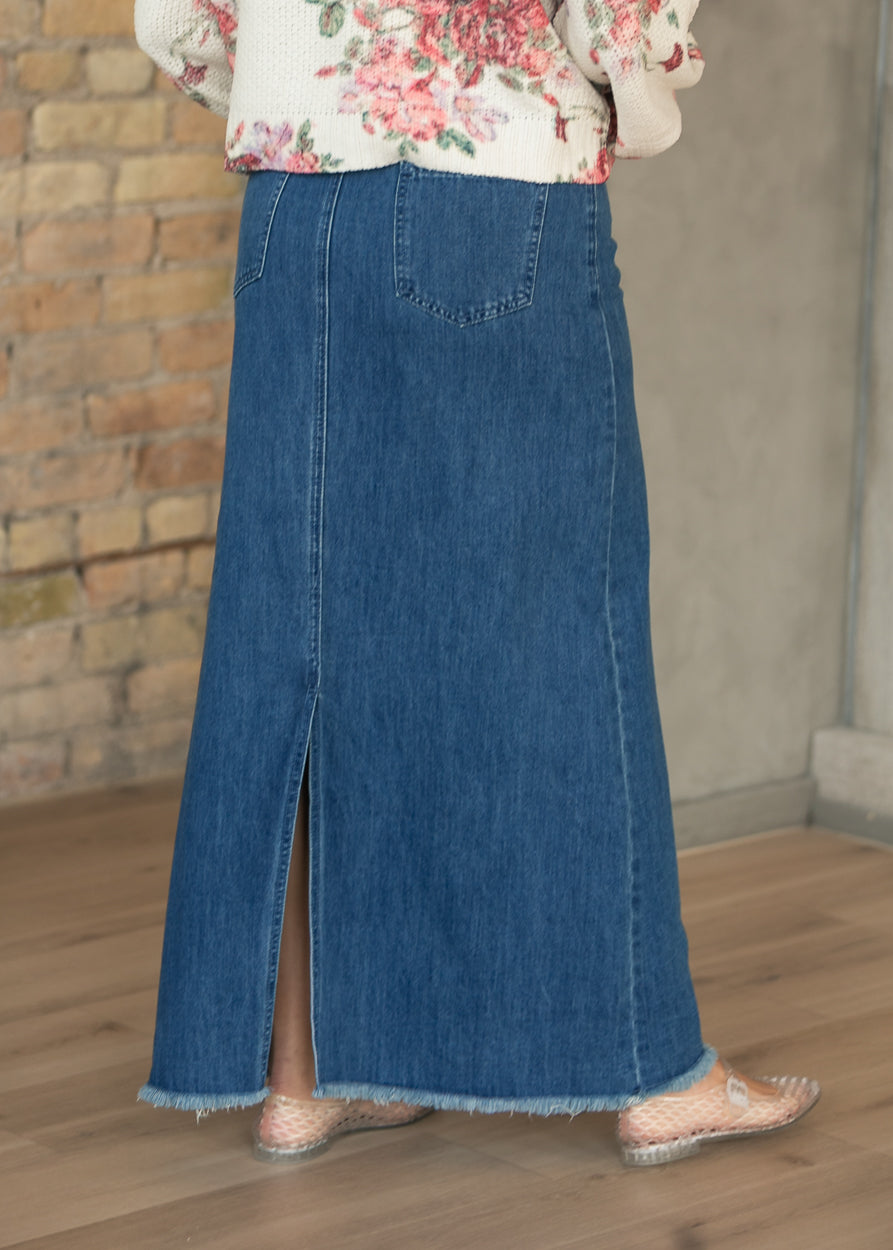 Back of medium-wash denim maxi skirt showing patch pockets, center back slit, and raw hem.