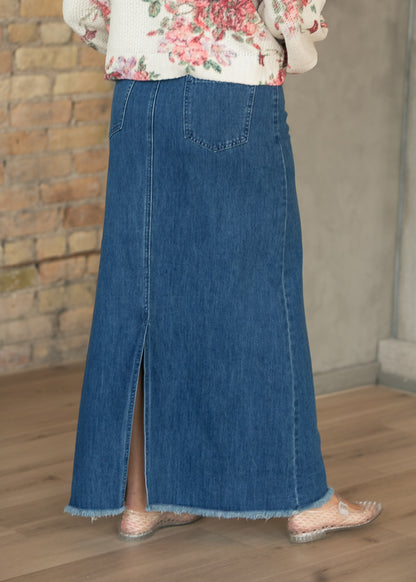 Back of medium-wash denim maxi skirt showing patch pockets, center back slit, and raw hem.