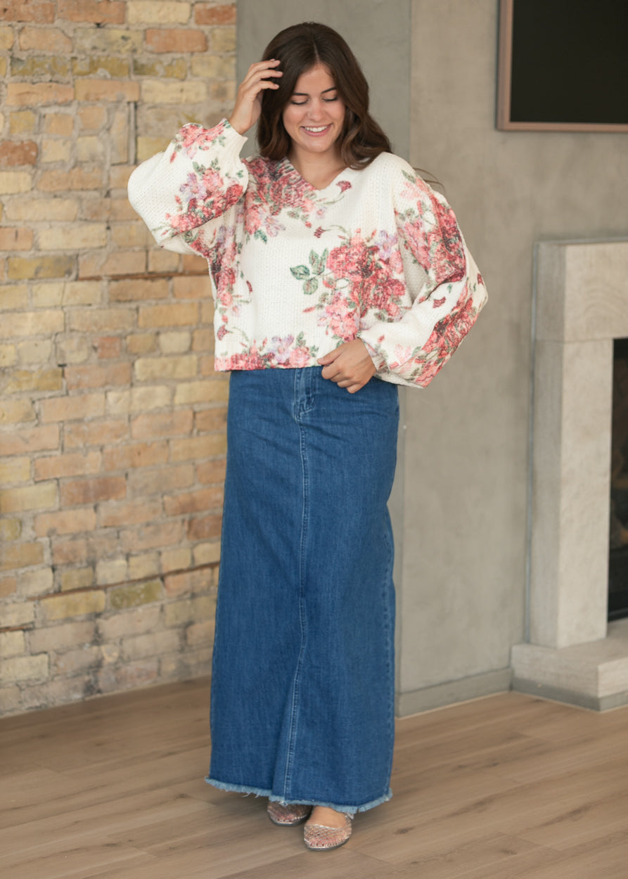 Medium-wash denim maxi skirt with raw hem and front pockets, styled with a floral sweater and flats.