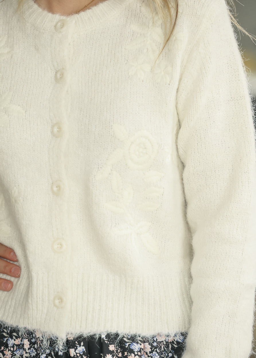 Close-up of ivory knit cardigan with tonal floral embroidery, functional buttons, and ribbed hem.