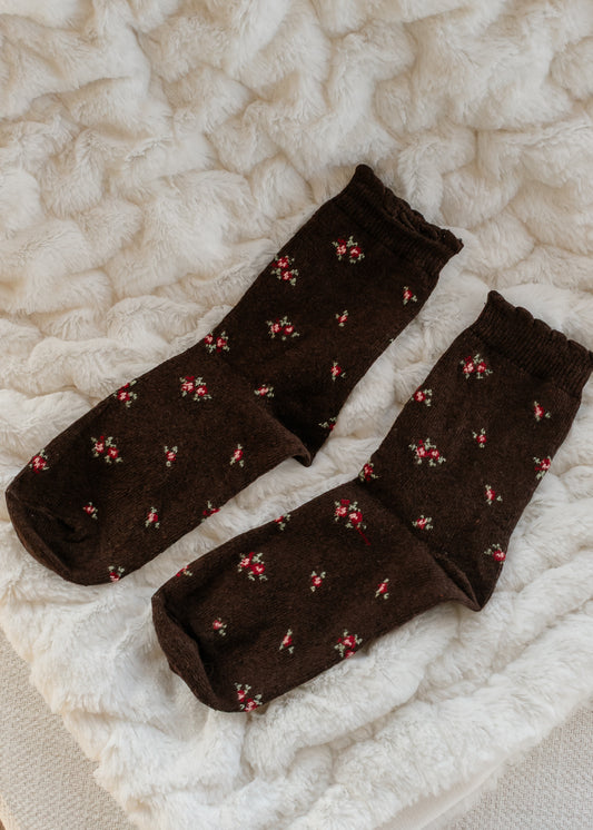 Brown cotton crew socks with tiny red and green floral print and ribbed cuffs, displayed on a soft cream textured blanket.