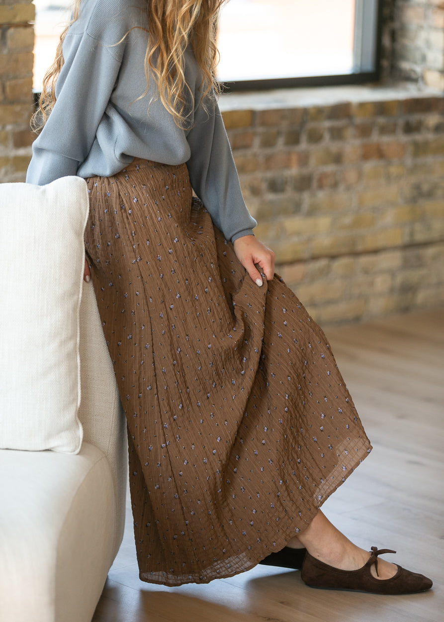 A woman wearing a brown pleated maxi skirt with a small blue and white pattern, styled with a light blue long-sleeve top and brown flat shoes, sitting on a white sofa in a room with a brick wall and window.