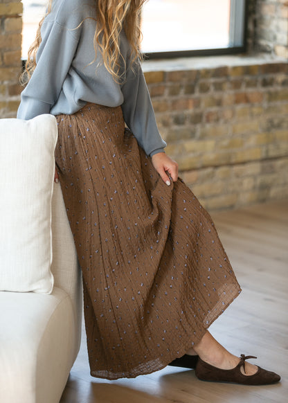A woman wearing a brown pleated maxi skirt with a small blue and white pattern, styled with a light blue long-sleeve top and brown flat shoes, sitting on a white sofa in a room with a brick wall and window.