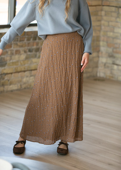 A person wearing a long pleated brown skirt with small scattered blue and white embroidered details, paired with a light blue sweatshirt and dark brown flats, standing in a room with a brick wall and wood floor.