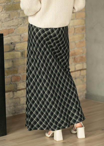 A person wearing a long black skirt with a white diagonal plaid pattern, paired with a cream knit sweater and white block-heel sandals, standing in front of a brick wall.