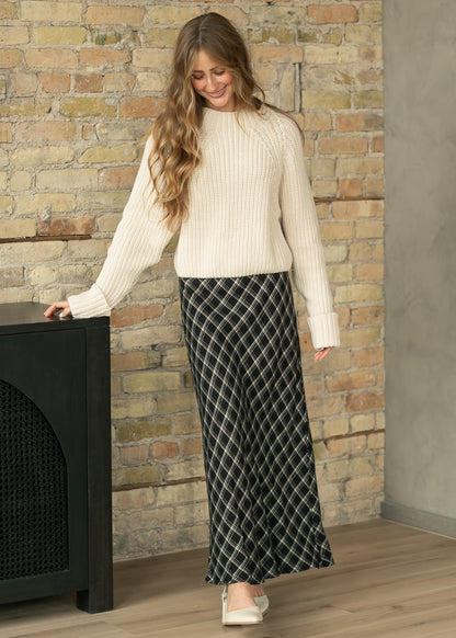 A woman wearing a cream ribbed knit mock-neck sweater and a black maxi skirt with a white geometric diamond pattern, standing in front of a brick wall.