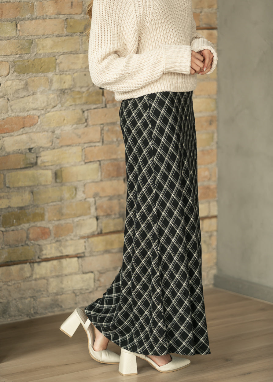 A person wearing a cream ribbed knit sweater and a black wide-leg skirt with a white diagonal diamond grid pattern, standing in front of a brick wall.