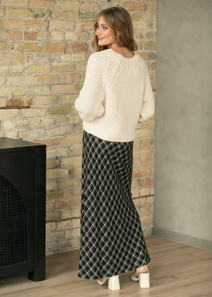 A woman wearing a cream cable-knit sweater and a black and white diamond-patterned maxi skirt, standing in front of a brick wall.