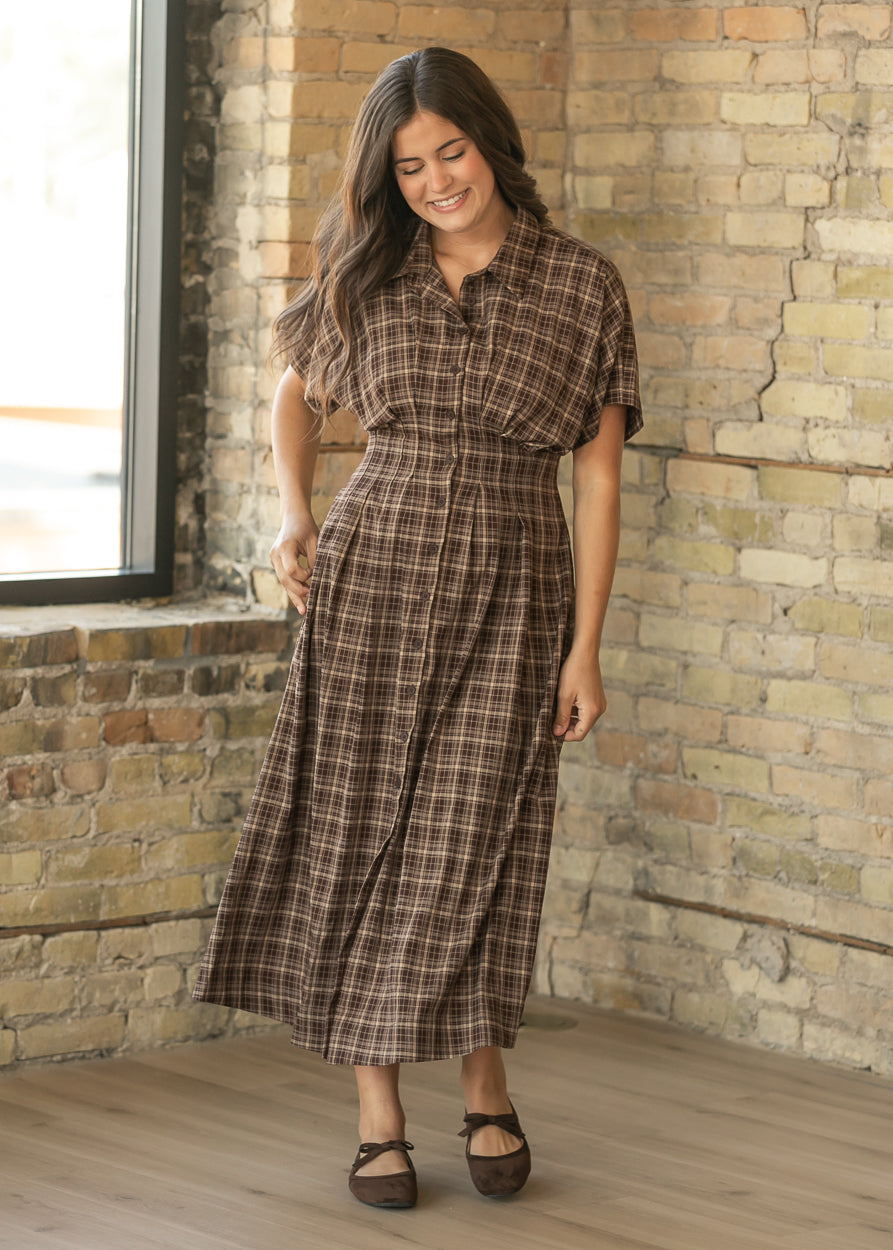 A woman wearing a brown and beige plaid midi shirt dress with short sleeves, standing in a room with a brick wall and wood floor.