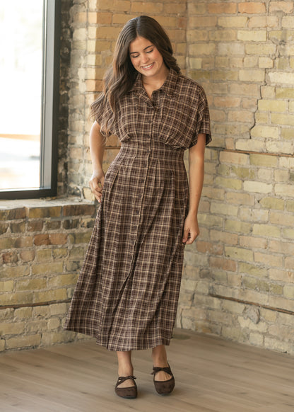 A woman wearing a brown and beige plaid midi shirt dress with short sleeves, standing in a room with a brick wall and wood floor.