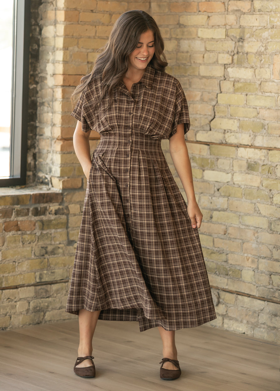 A woman wearing a brown plaid midi shirt dress with short sleeves and a button-down front, standing in front of a rustic brick wall.