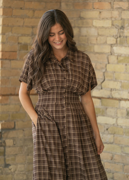 A woman wearing a brown and beige plaid shirt dress, standing in front of a rustic brick wall.