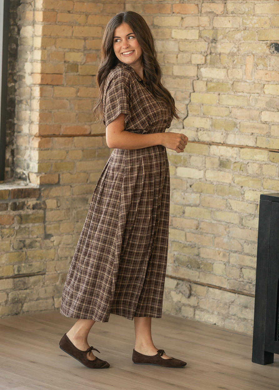 A woman wearing a brown and beige plaid midi dress with short sleeves and brown flat shoes, standing in front of a brick wall.