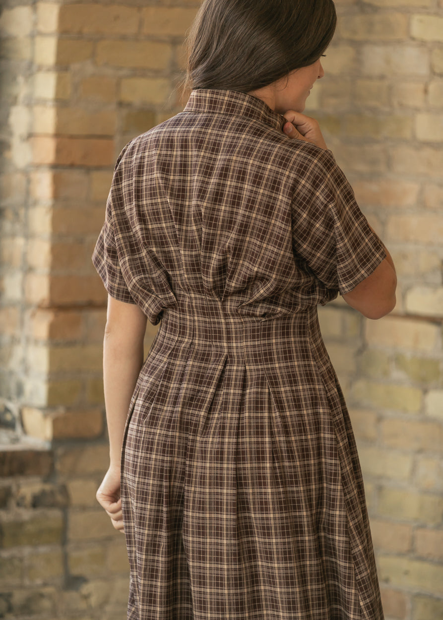 A woman wearing a brown and beige plaid short-sleeve dress, shown from behind against a rustic brick wall.