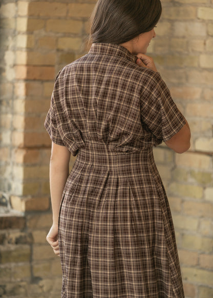 A woman wearing a brown and beige plaid short-sleeve dress, shown from behind against a rustic brick wall.