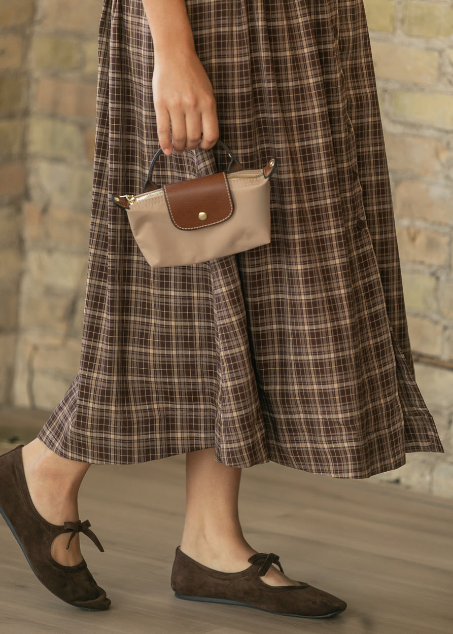 A person wearing a brown plaid midi skirt and dark brown suede ballet flats, holding a beige and brown handbag against a rustic brick wall background.