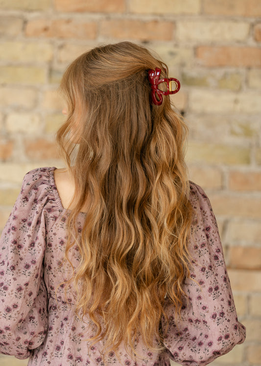 AK glossy burgundy bow hair claw clip styled in long wavy hair with a floral dress.