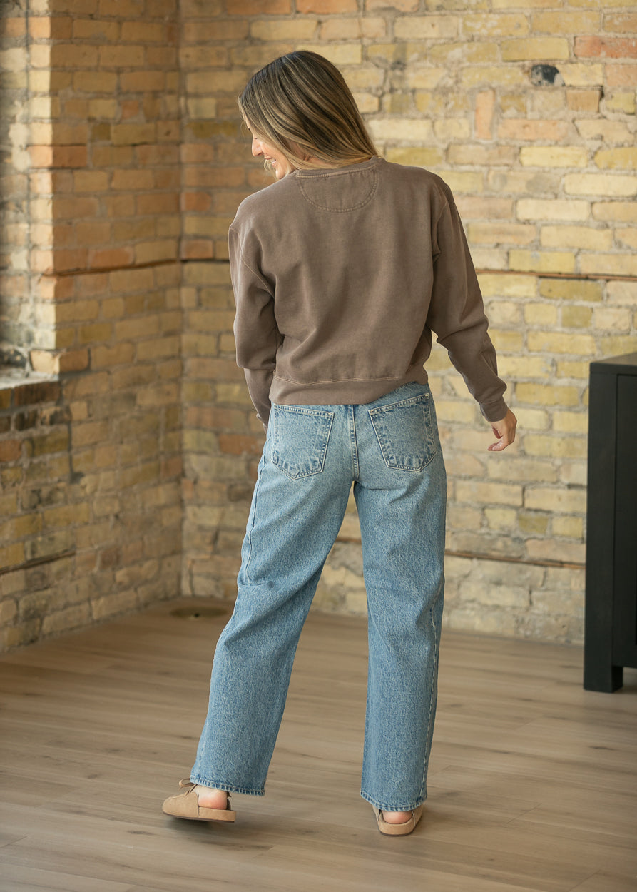A woman is shown from behind wearing a brown crewneck sweatshirt and light-wash wide-leg jeans, standing in a room with a brick wall and wooden floor.