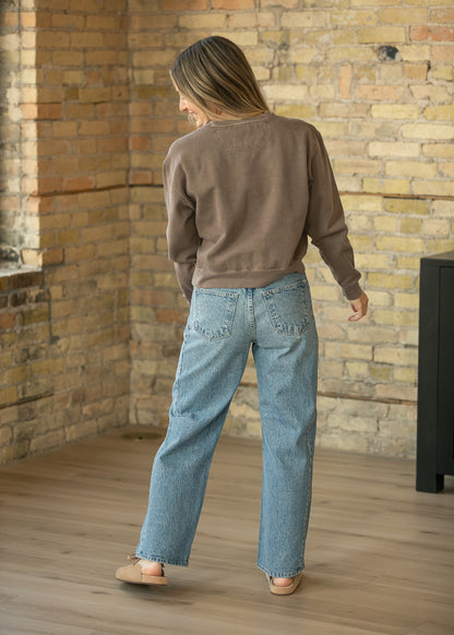 A woman is shown from behind wearing a brown crewneck sweatshirt and light-wash wide-leg jeans, standing in a room with a brick wall and wooden floor.