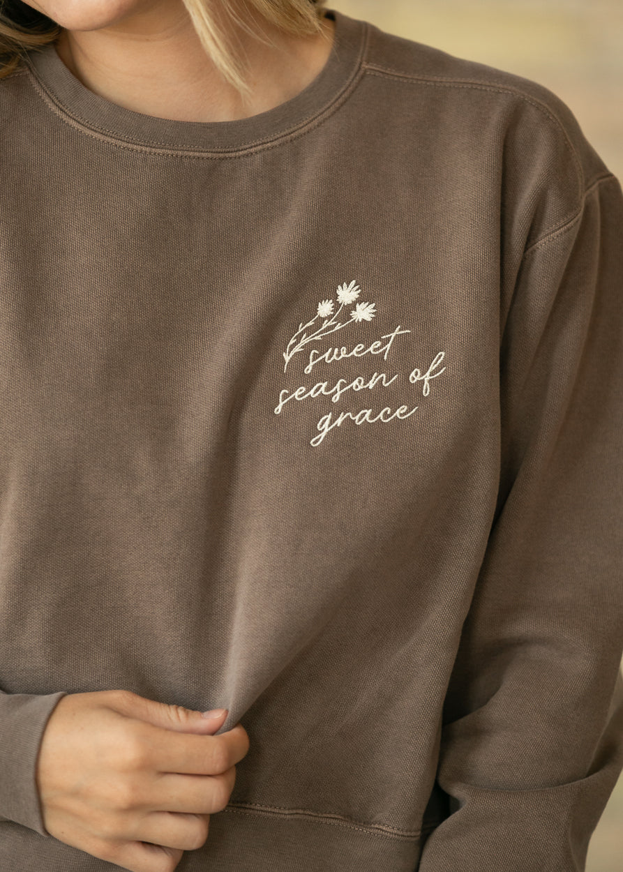 A close-up of a person wearing a brown crewneck sweatshirt with white embroidered text reading 'sweet season of grace' and a small floral design on the left chest.