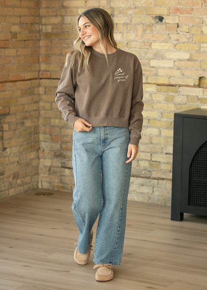 A woman wearing a brown crewneck sweatshirt with a small white graphic on the left chest, paired with light blue wide-leg jeans and beige shoes, standing in front of a brick wall.
