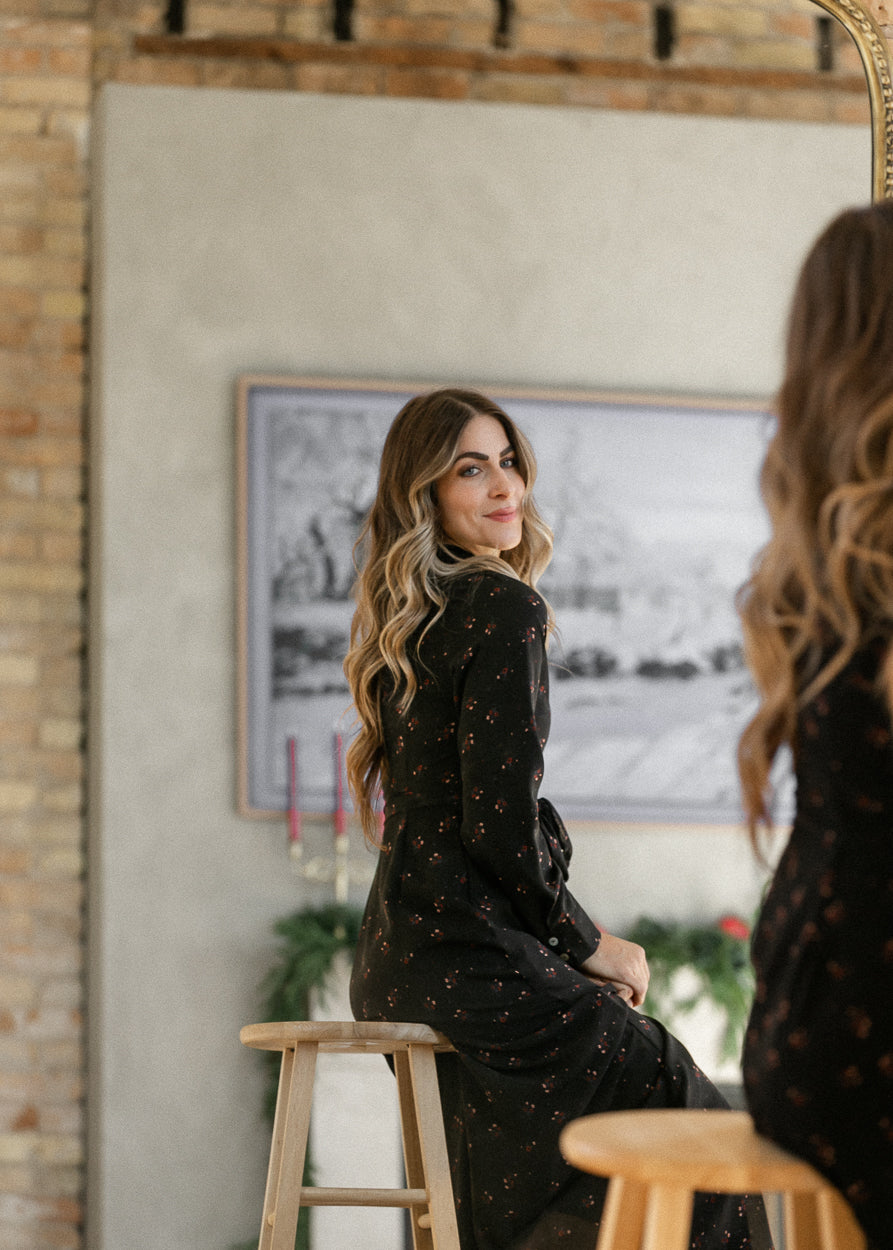 Gracelyn black floral maxi dress shown in a mirror reflection, worn seated on a wooden stool.