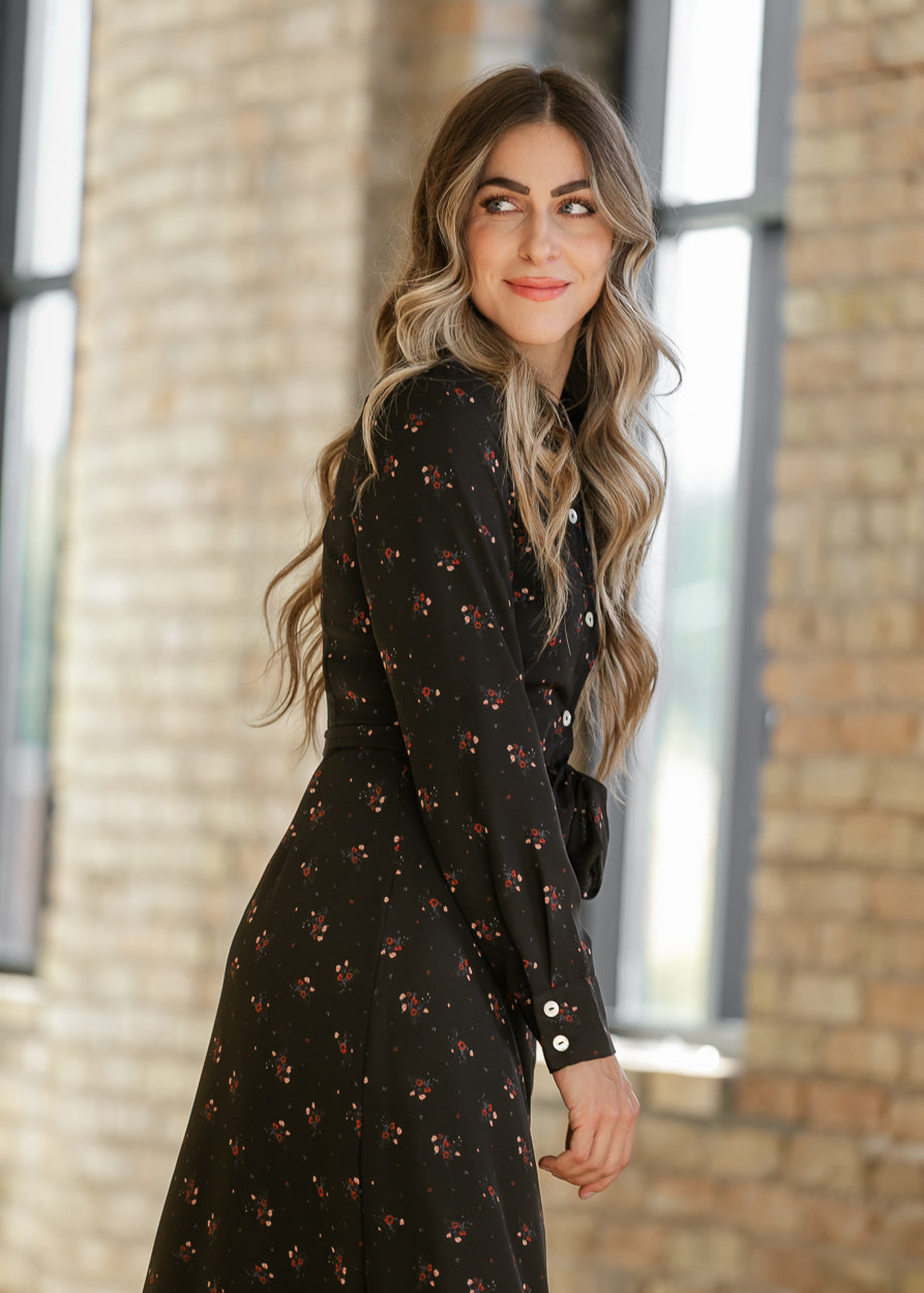 Black floral dress shown at an angle, highlighting long sleeves, contrast cuff buttons, and a matching waist tie.