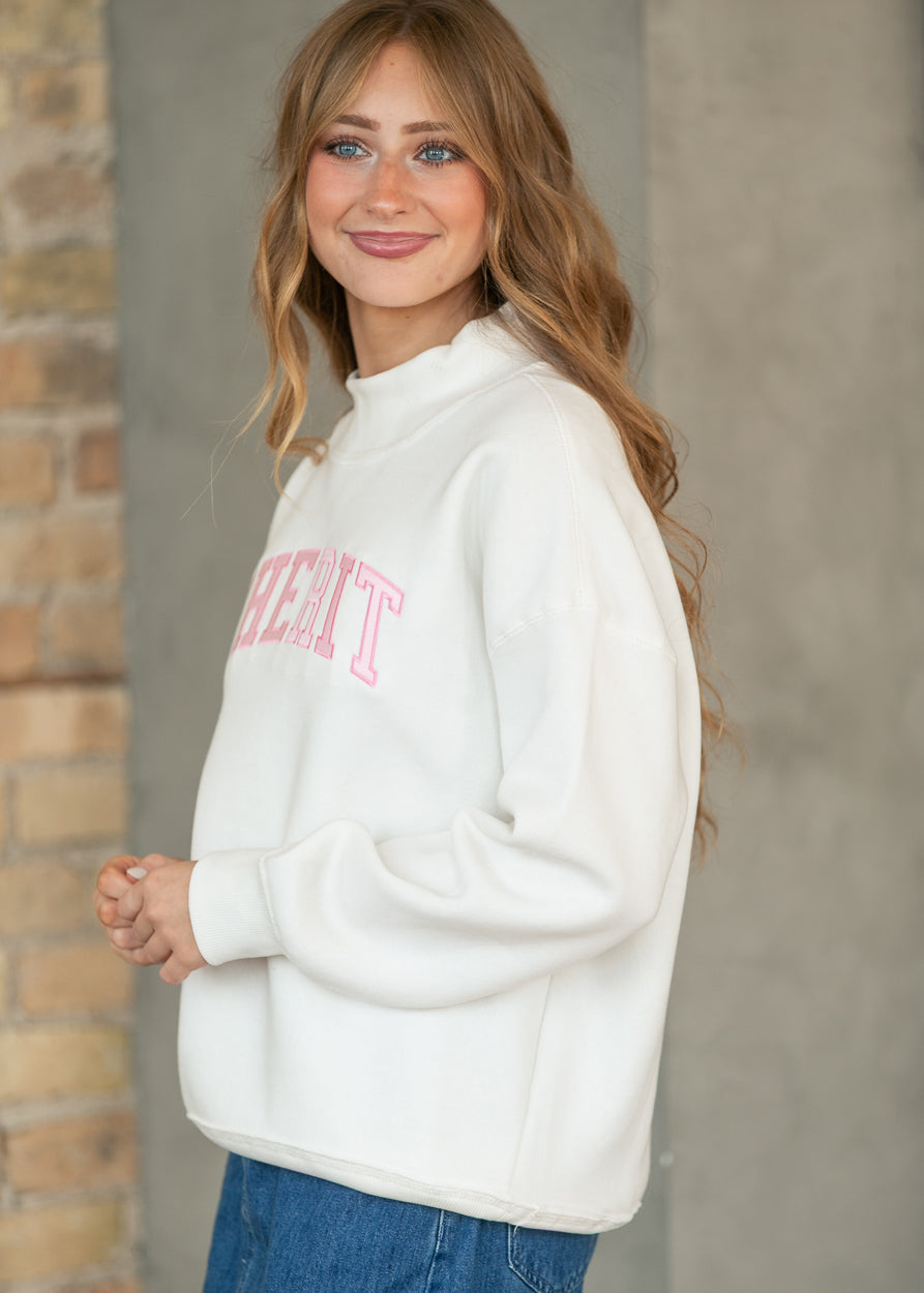 A woman wearing a white mock-neck sweatshirt with pink collegiate-style lettering on the chest, paired with blue jeans, standing against a gray concrete wall and brick column.
