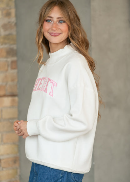 A woman wearing a white mock-neck sweatshirt with pink collegiate-style lettering on the chest, paired with blue jeans, standing against a gray concrete wall and brick column.