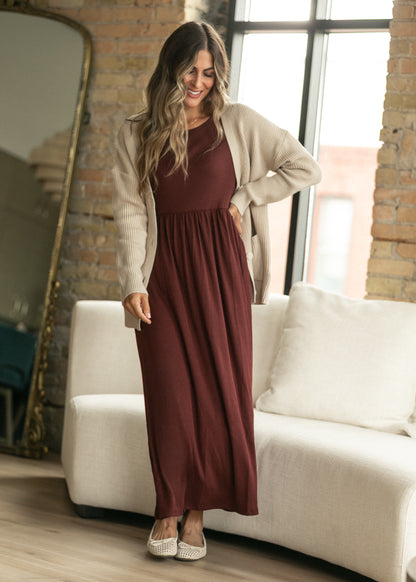 A woman models a long-sleeve burgundy maxi dress with a cream cardigan in a bright, modern loft setting with a white sofa and brick walls.