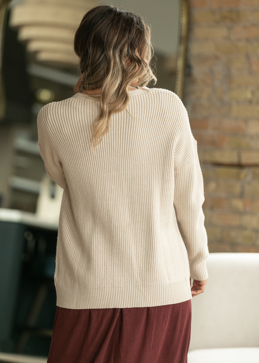 A woman viewed from behind wearing a cream ribbed knit sweater and a burgundy skirt in an indoor setting with a brick wall background.