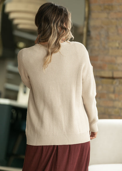 A woman viewed from behind wearing a cream ribbed knit sweater and a burgundy skirt in an indoor setting with a brick wall background.