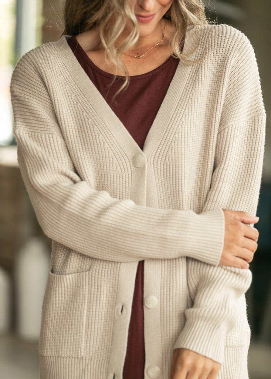 A woman wearing a cream ribbed cardigan over a burgundy top, shown from the shoulders to the hips in a softly blurred indoor setting.