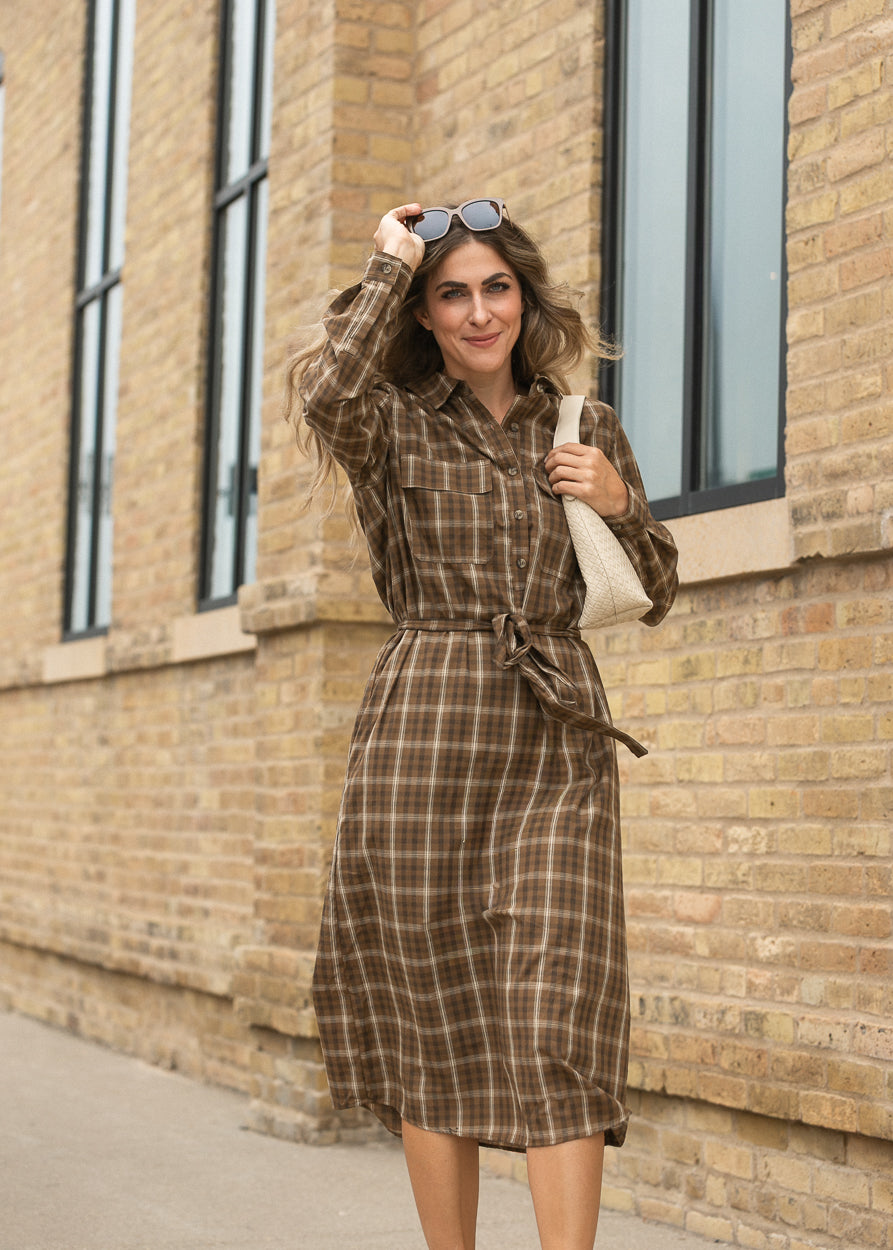 Brown plaid midi dress with long sleeves and defined tie waist, photographed beside a brick building.