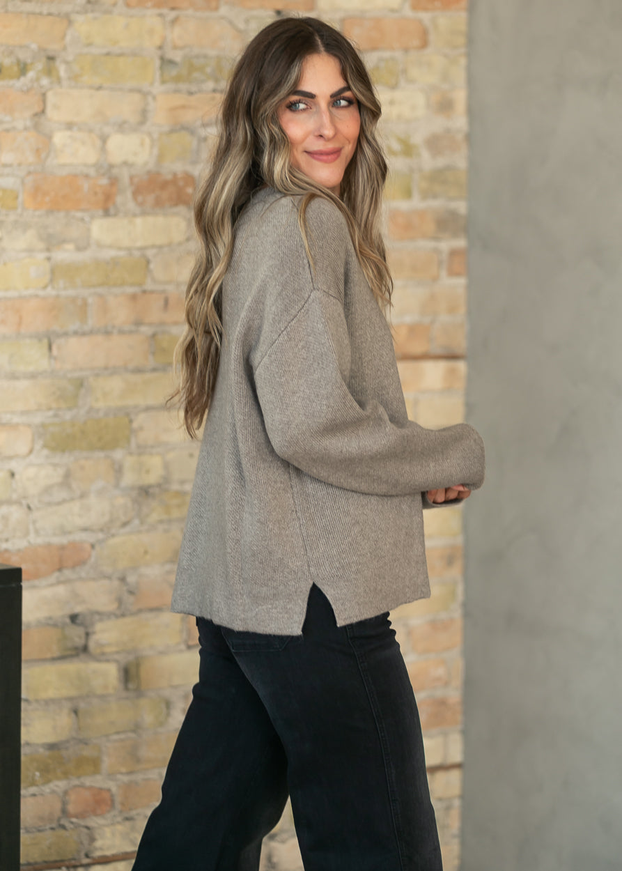 A woman with long wavy brown hair is shown from the back and side, wearing a light taupe-gray knit sweater and black pants, standing in front of a brick wall.