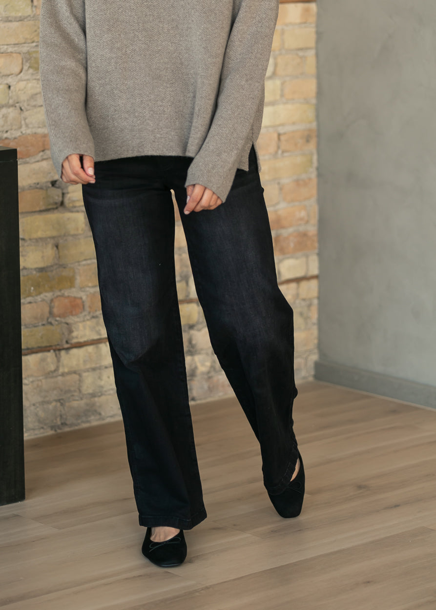 A person wearing dark wide-leg jeans, a light beige knit sweater, and black flats, standing on a light wood floor in front of a brick wall.