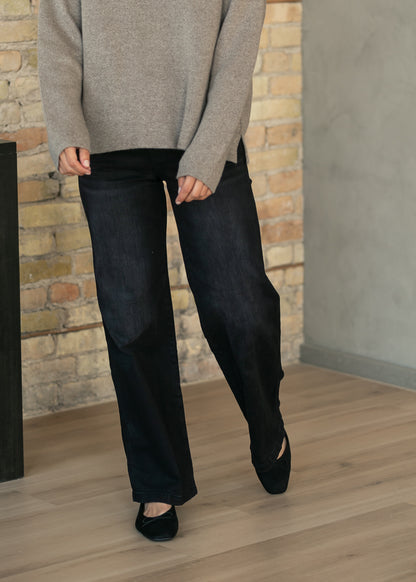 A person wearing dark wide-leg jeans, a light beige knit sweater, and black flats, standing on a light wood floor in front of a brick wall.