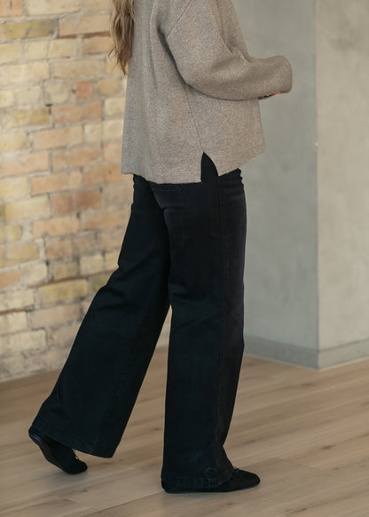 A person wearing wide-leg black pants and a light beige knit sweater, standing in a room with a brick wall and wood floor.
