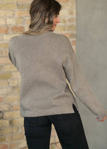 A woman wearing a light taupe knit sweater and dark jeans, shown from the back against a split brick and concrete wall background.